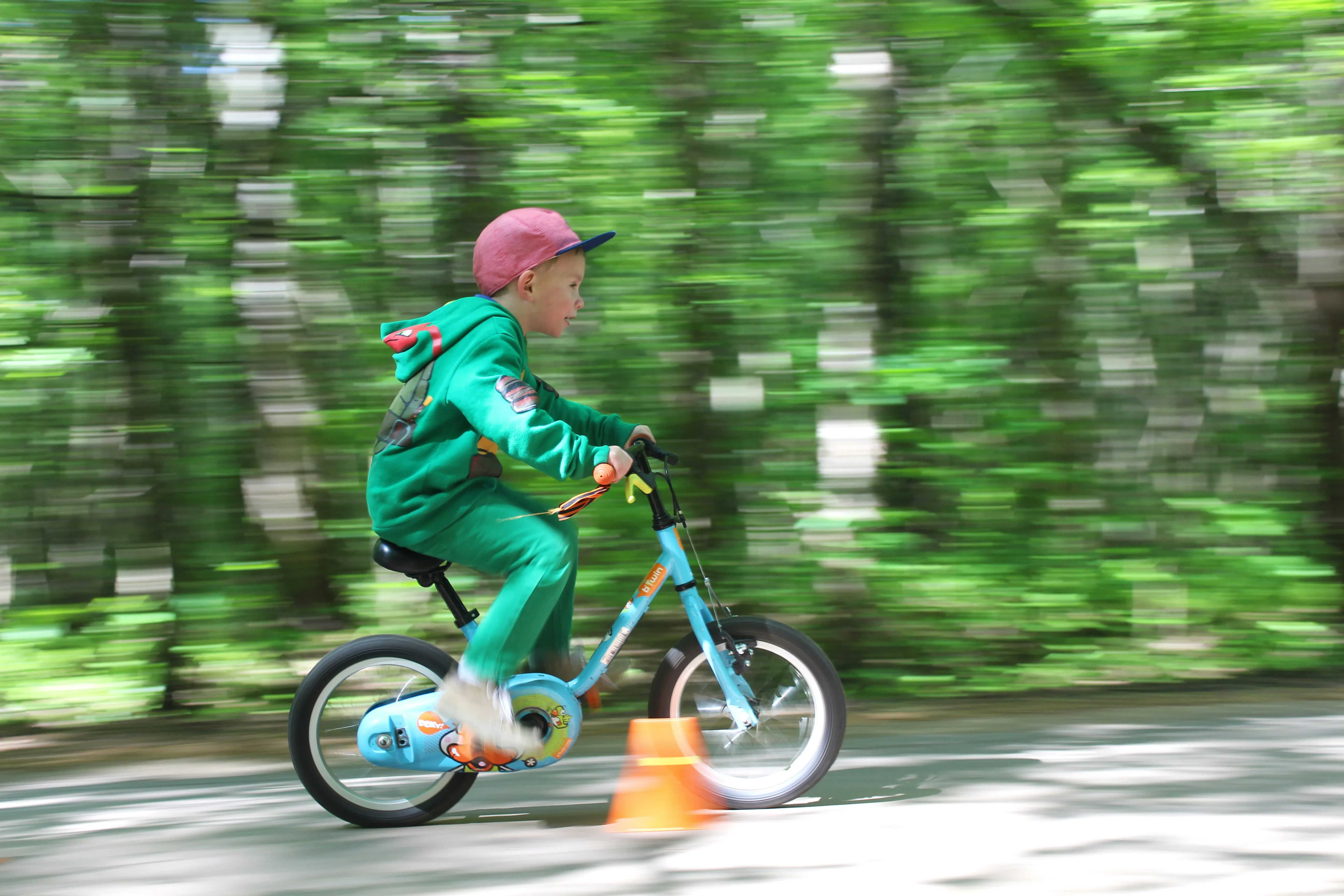 A young child in a green tracksuit riding a bike fast along a tree-lined path, the background blurred with motion